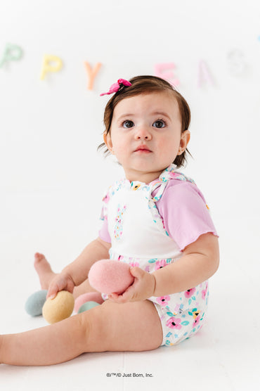 Baby sitting on a white surface holding a pink toy, wearing a PEEPS® romper with a lettuce hem lavender shirt underneath. 