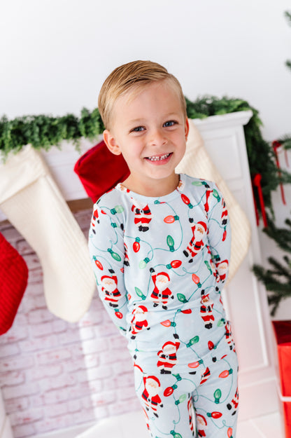A young boy smiling and wearing a light blue Christmas-themed long-sleeve bamboo pajama set with a Santa Claus and holiday light print, posed in front of a decorated fireplace.