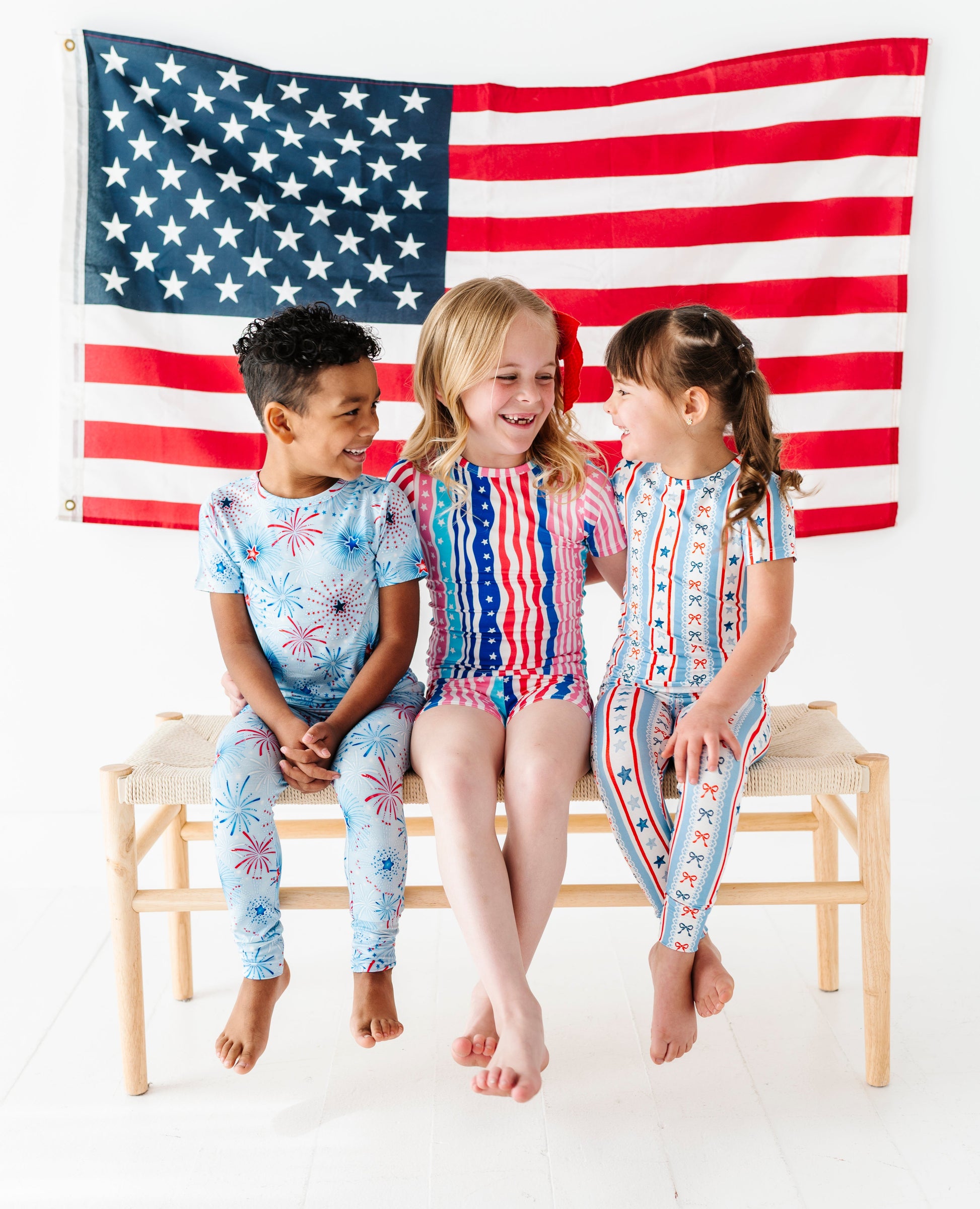 Three children in patriotic bamboo two-piece pajamas sitting on a bench with an American flag in the background