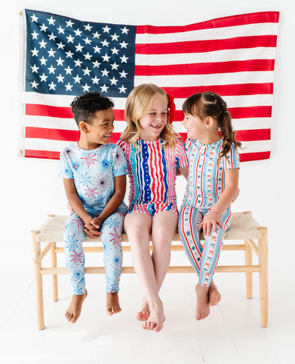 Three children in patriotic bamboo two-piece pajamas sitting on a bench with an American flag in the background
