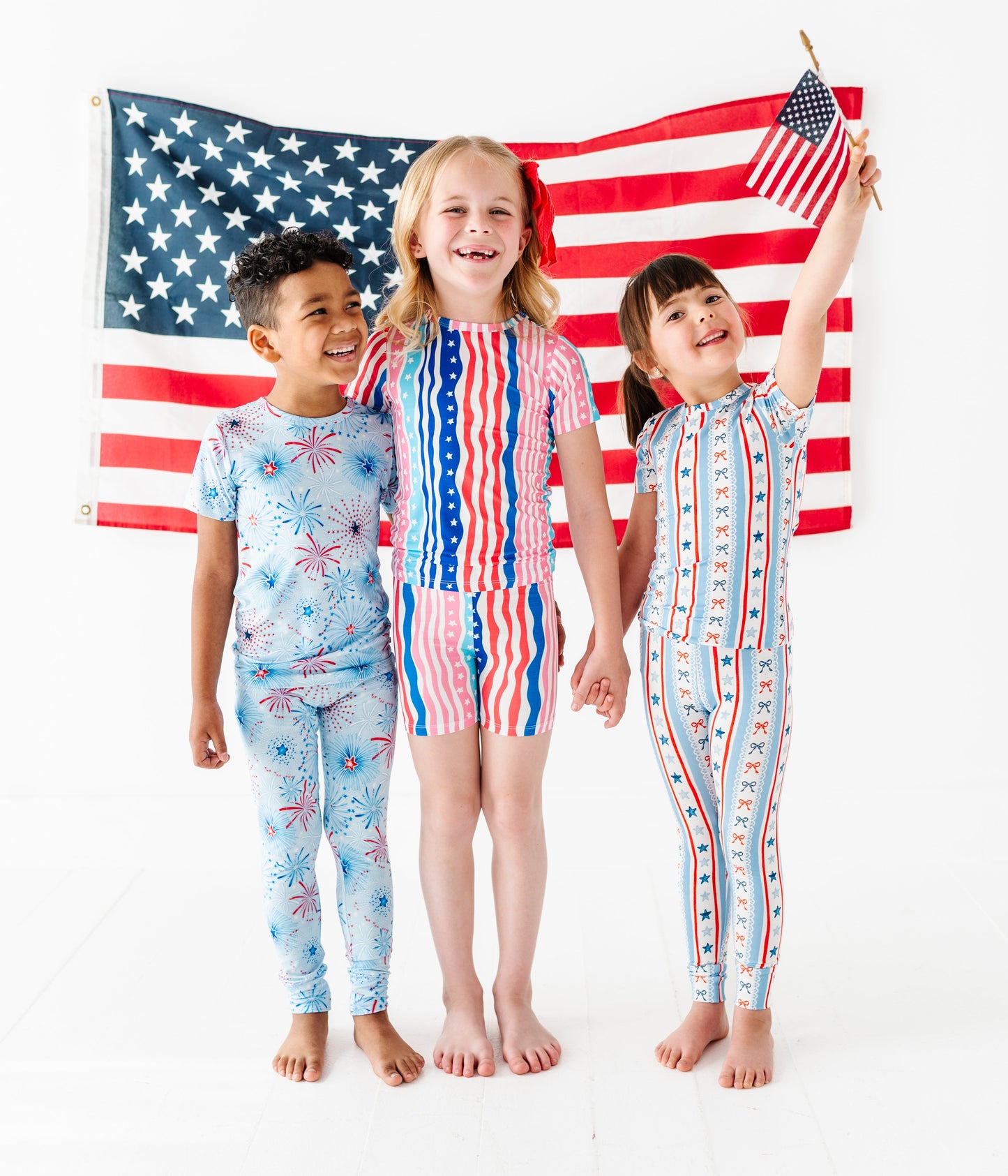 Three children in patriotic-themed bamboo pajamas holding an American flag on a white background