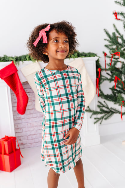 A little girl wearing a bamboo nightgown in a classic Christmas plaid print, standing in front of a fireplace with stockings and a decorated Christmas tree