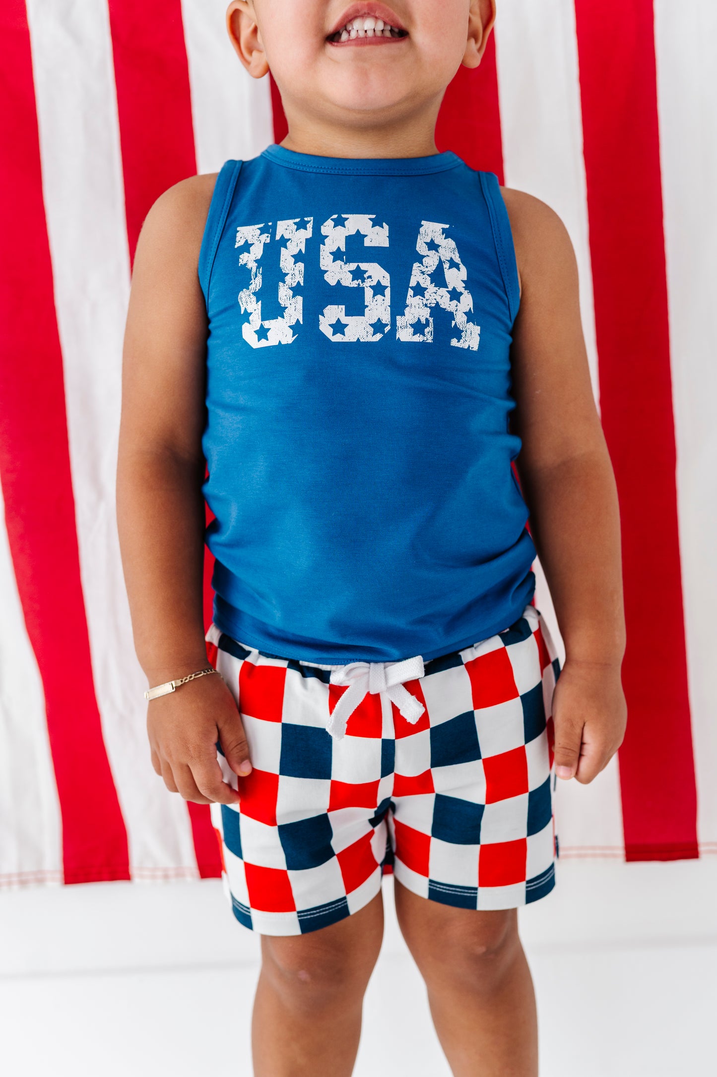 Child wearing a cobalt blue 'USA' bamboo tank top and red, white, and blue checkered shorts in front of an American flag.