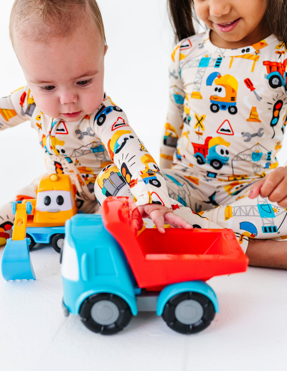 Two toddlers in matching construction-themed pajamas play with a blue and red toy dump truck on a white floor.