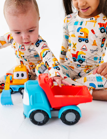 Two toddlers in matching construction-themed pajamas play with a blue and red toy dump truck on a white floor.