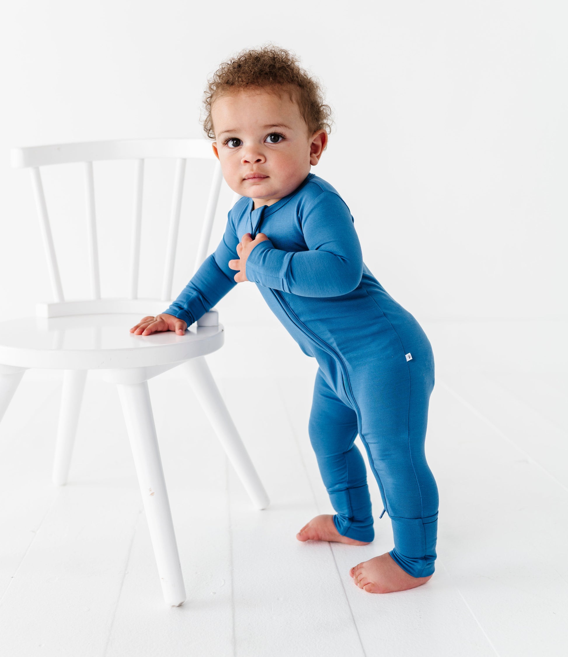 Baby in a blue bamboo convertible romper standing next to a white chair on a white background