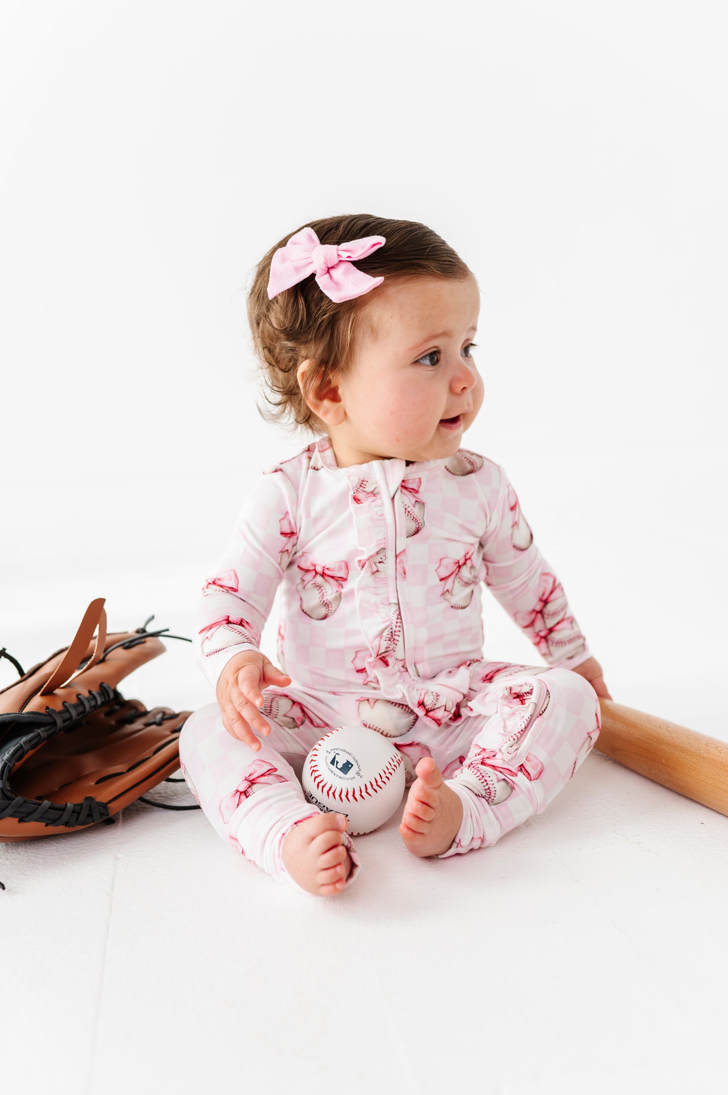 Baby in a pink bamboo outfit with a baseball design, sitting on a white surface.