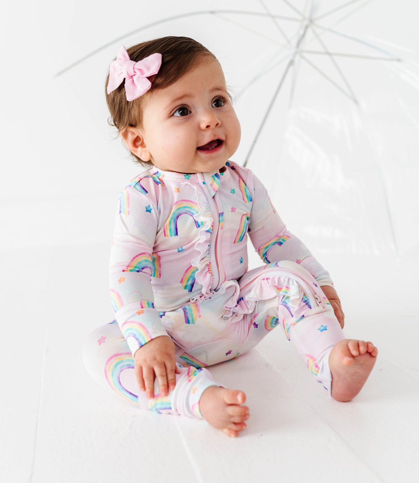 Baby wearing a rainbow bamboo onesie sitting on a white surface with an umbrella in the background