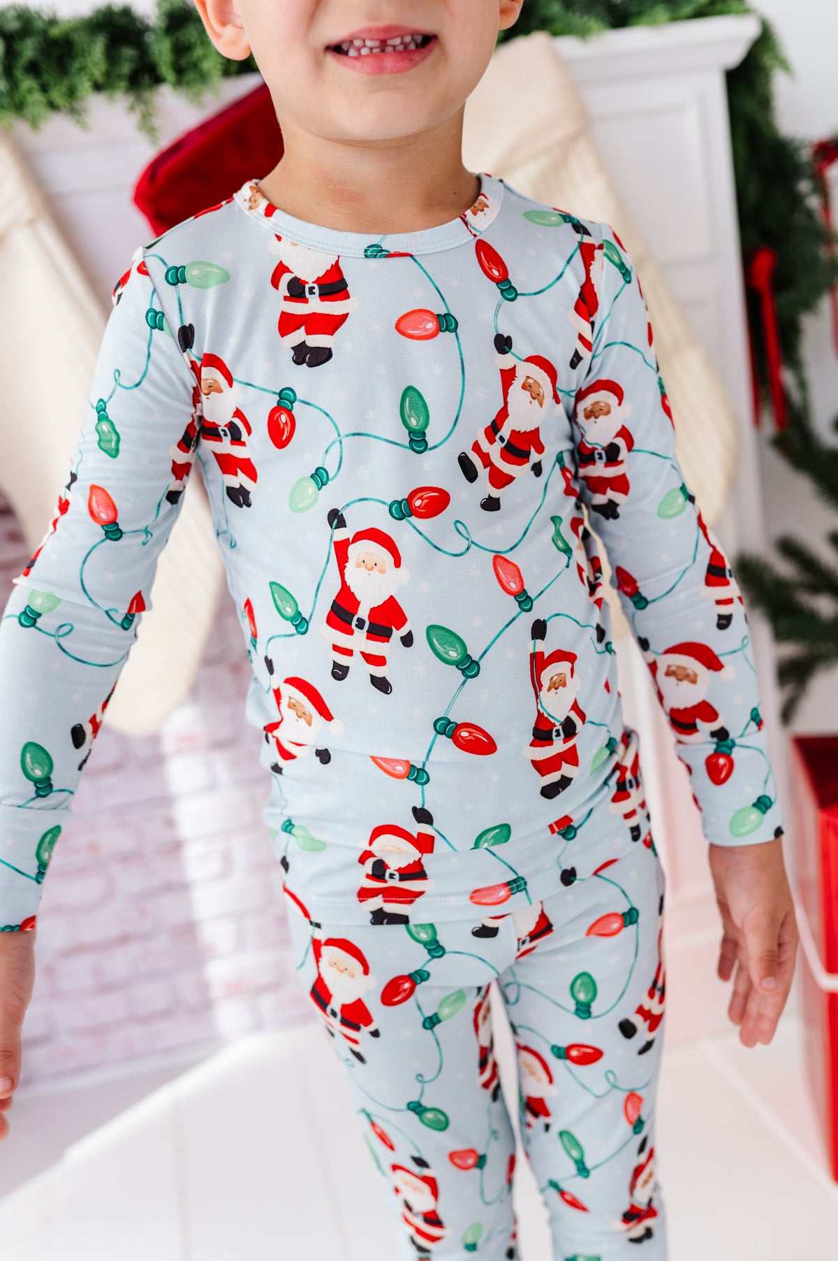 A child wearing a long-sleeve pajama top and matching pants set with a festive Christmas lights print featuring Santa Claus figures on a light blue background.