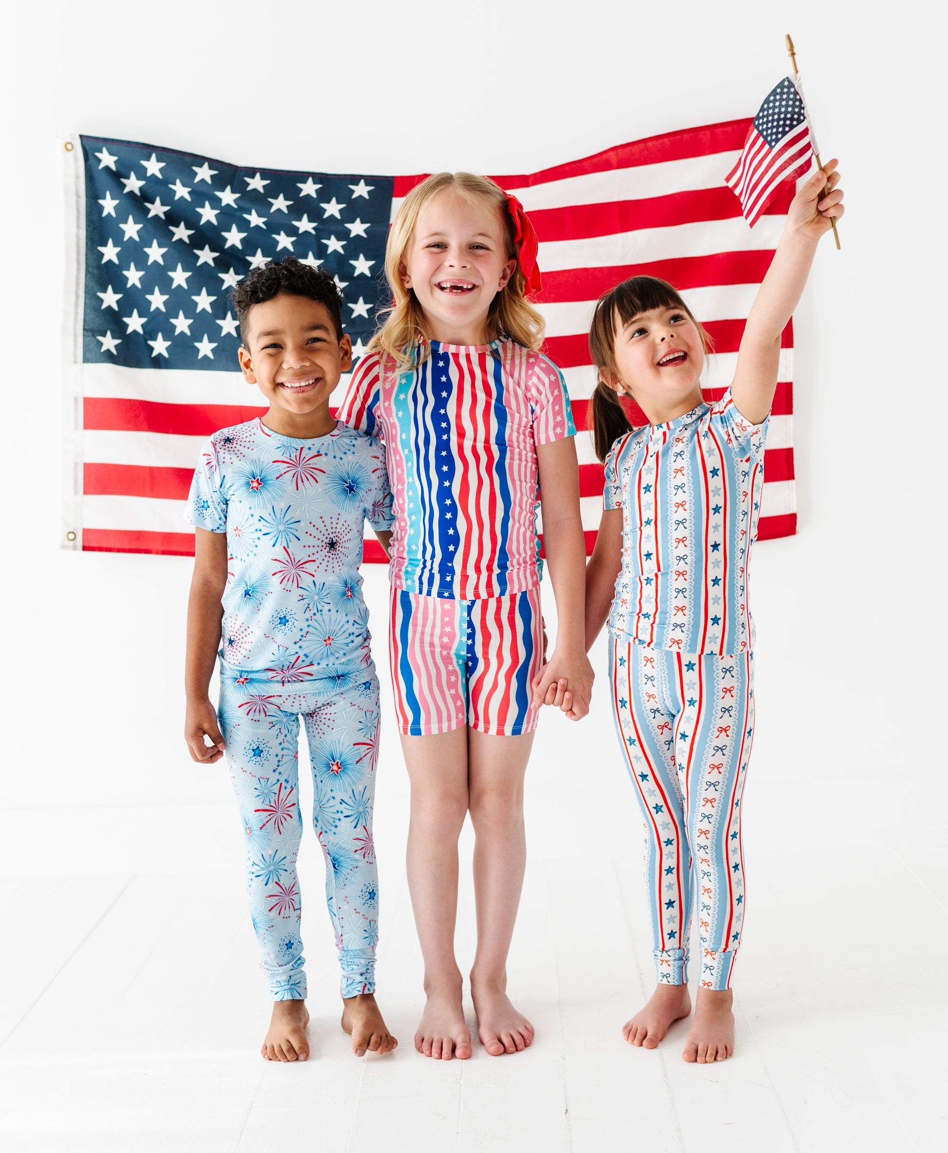 Three children wearing matching American flag-themed bamboo pajamas standing in front of an American flag.