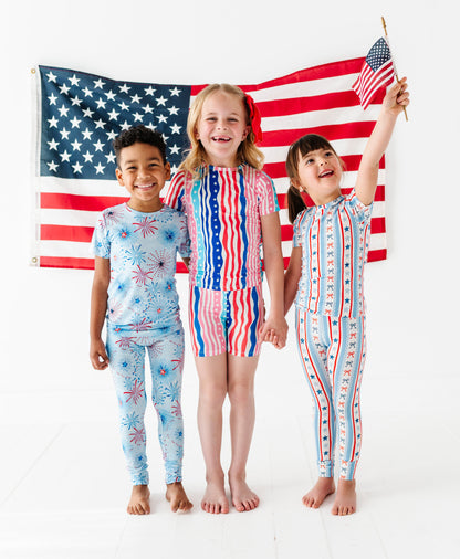 Three children wearing matching American flag-themed bamboo pajamas standing in front of an American flag.