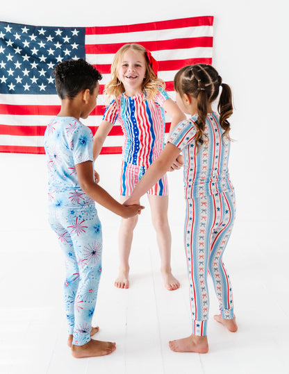 Three children in patriotic bamboo pajamas standing in front of an American flag.