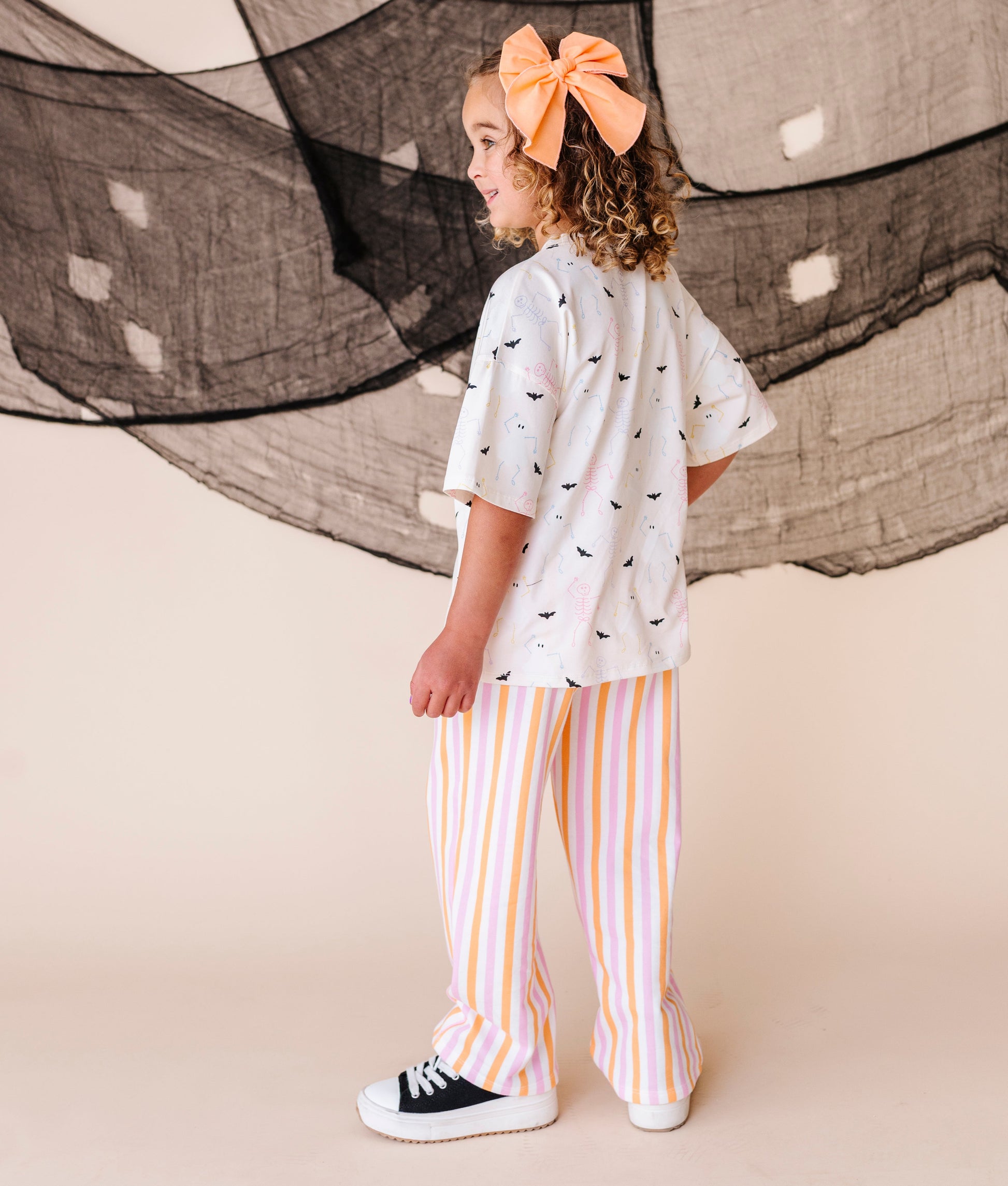 Child wearing a cream shirt with halloween patterns and striped pants, standing in front of large netting.