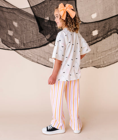 Child wearing a cream shirt with halloween patterns and striped pants, standing in front of large netting.