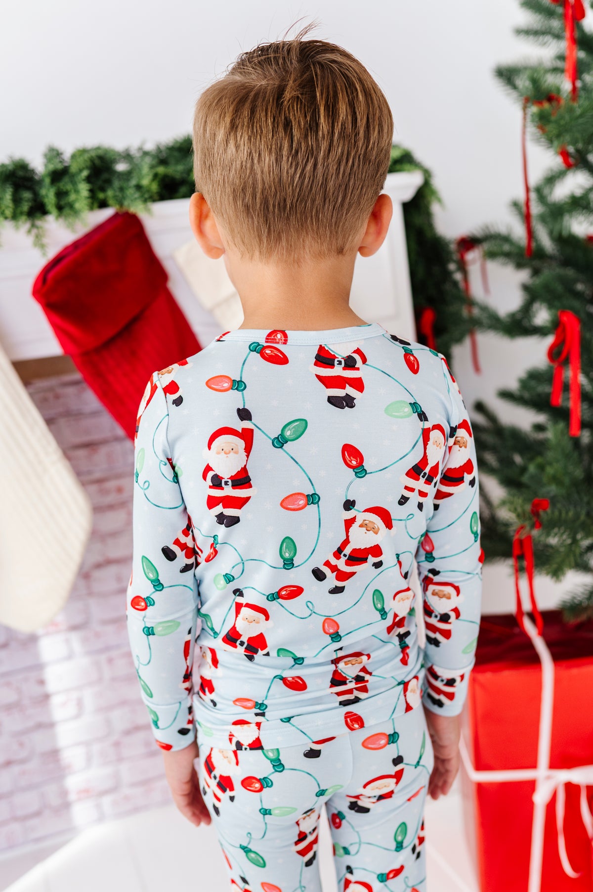 A child viewed from behind wearing a light blue long-sleeve bamboo pajama set with a Santa Claus and Christmas light string pattern, standing in a holiday-themed room with a Christmas tree and stockings in the background.