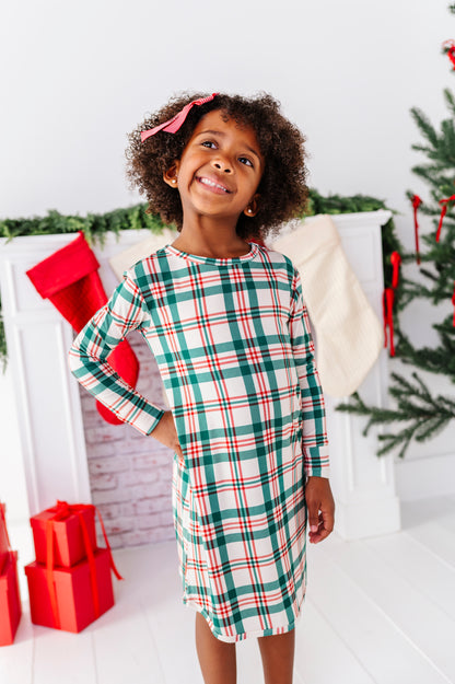 A little girl wearing a bamboo nightgown in a classic Christmas plaid print, standing in front of a fireplace with stockings and a decorated Christmas tree