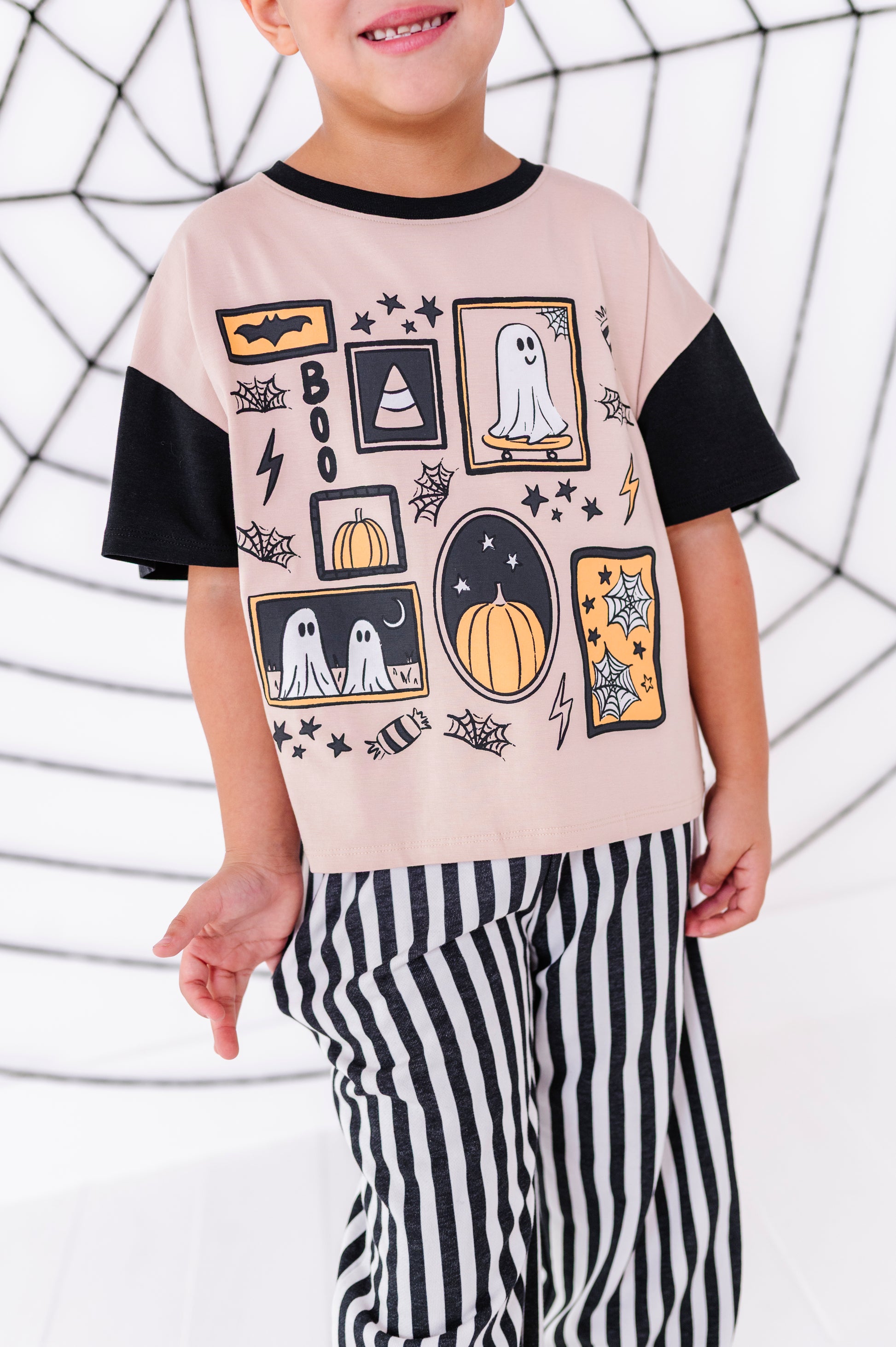 A child wearing a light pink short-sleeve T-shirt with black sleeves and a black crew neck, decorated with a Halloween-themed graphic print featuring ghosts, pumpkins, bats, and stars. The child is also wearing black and white vertically striped pants, posed against a white background with a black spiderweb pattern.