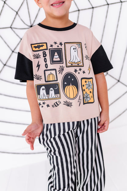 A child wearing a light pink short-sleeve T-shirt with black sleeves and a black crew neck, decorated with a Halloween-themed graphic print featuring ghosts, pumpkins, bats, and stars. The child is also wearing black and white vertically striped pants, posed against a white background with a black spiderweb pattern.