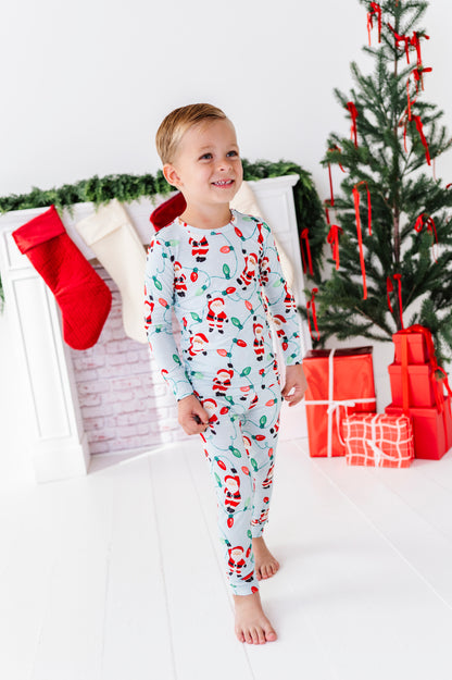 A young boy wearing a light blue long-sleeve bamboo pajama set with a Santa Claus pattern, standing in a festive holiday room with a Christmas tree, stockings, and wrapped gifts.