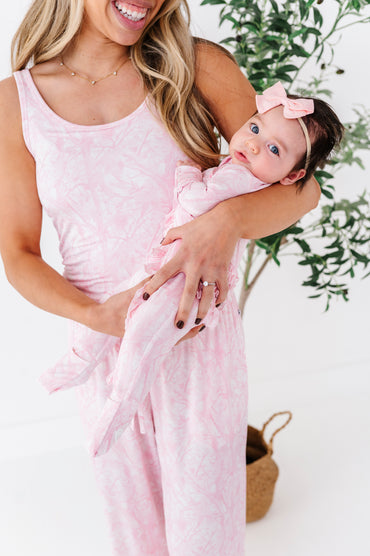 Woman in a pink tree camo  lounge set holding a baby also in pink camo, with a white background and greenery.