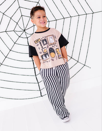 A young boy modeling a Halloween-themed t-shirt and striped pants in front of a large black spiderweb backdrop on a white studio floor.