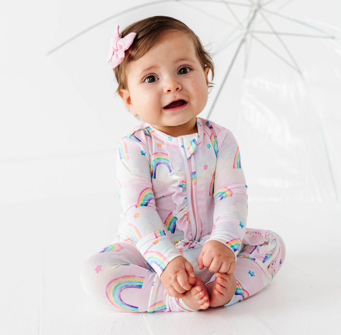 Baby wearing a colorful bamboo zip romper with watercolor rainbow patterns, sitting under an umbrella on a white background.