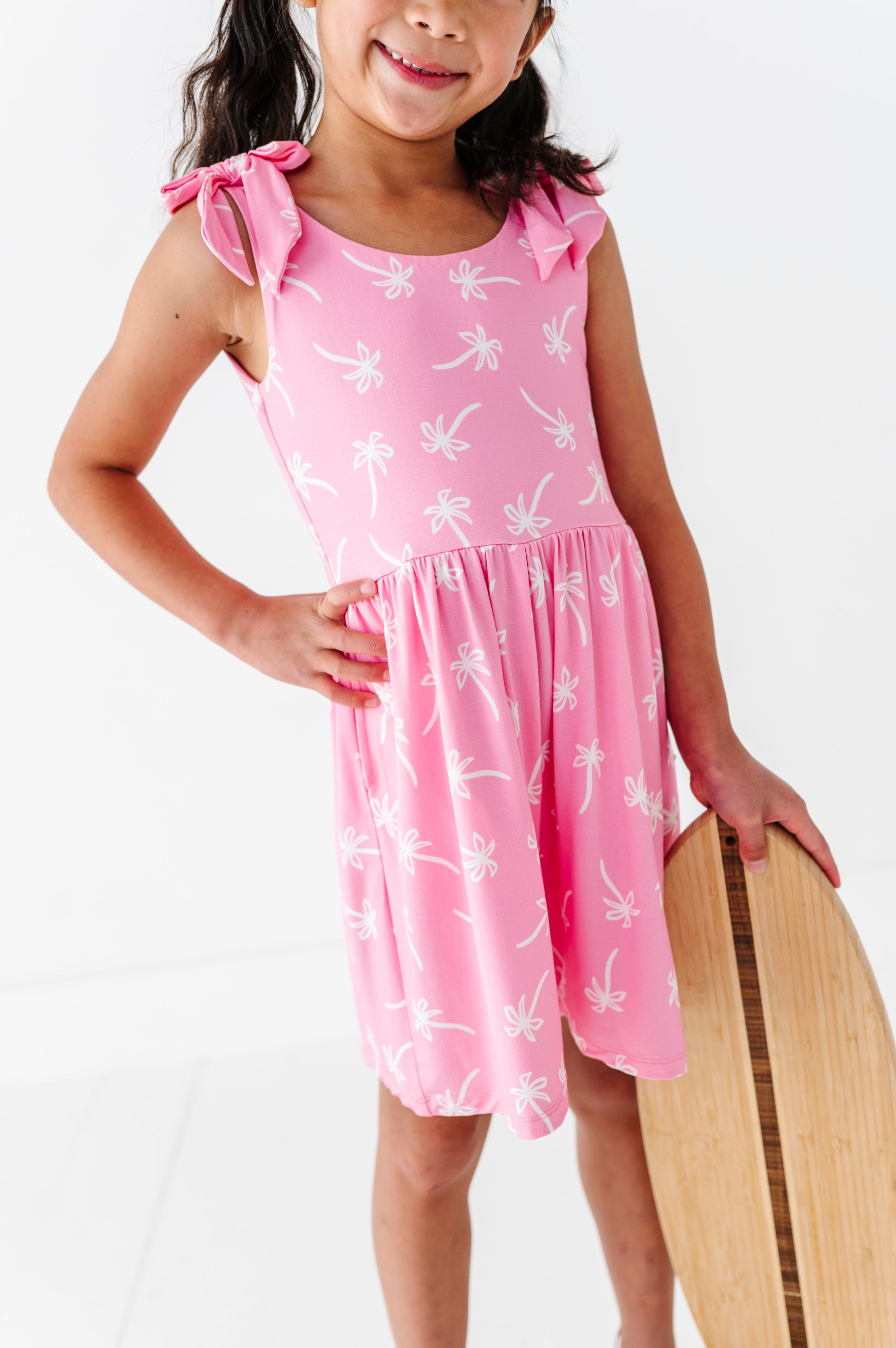 Young girl wearing a pink romper with white patterns, holding a wooden surfboard on a white background