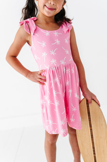 Young girl wearing a pink romper with white patterns, holding a wooden surfboard on a white background