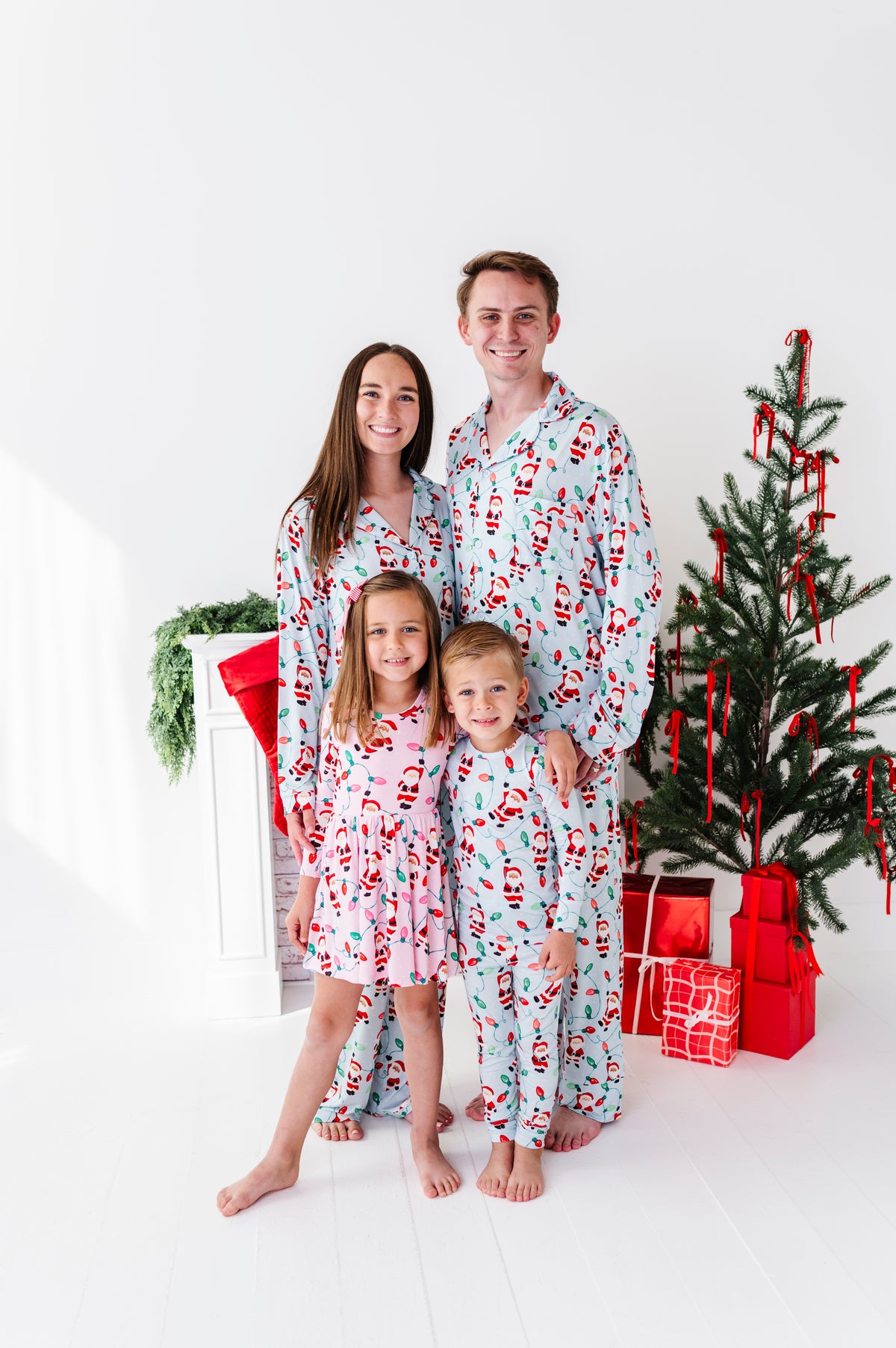 A family of four wearing matching Santa Claus print bamboo pajamas stands together in a bright white room next to a decorated Christmas tree and wrapped gifts.