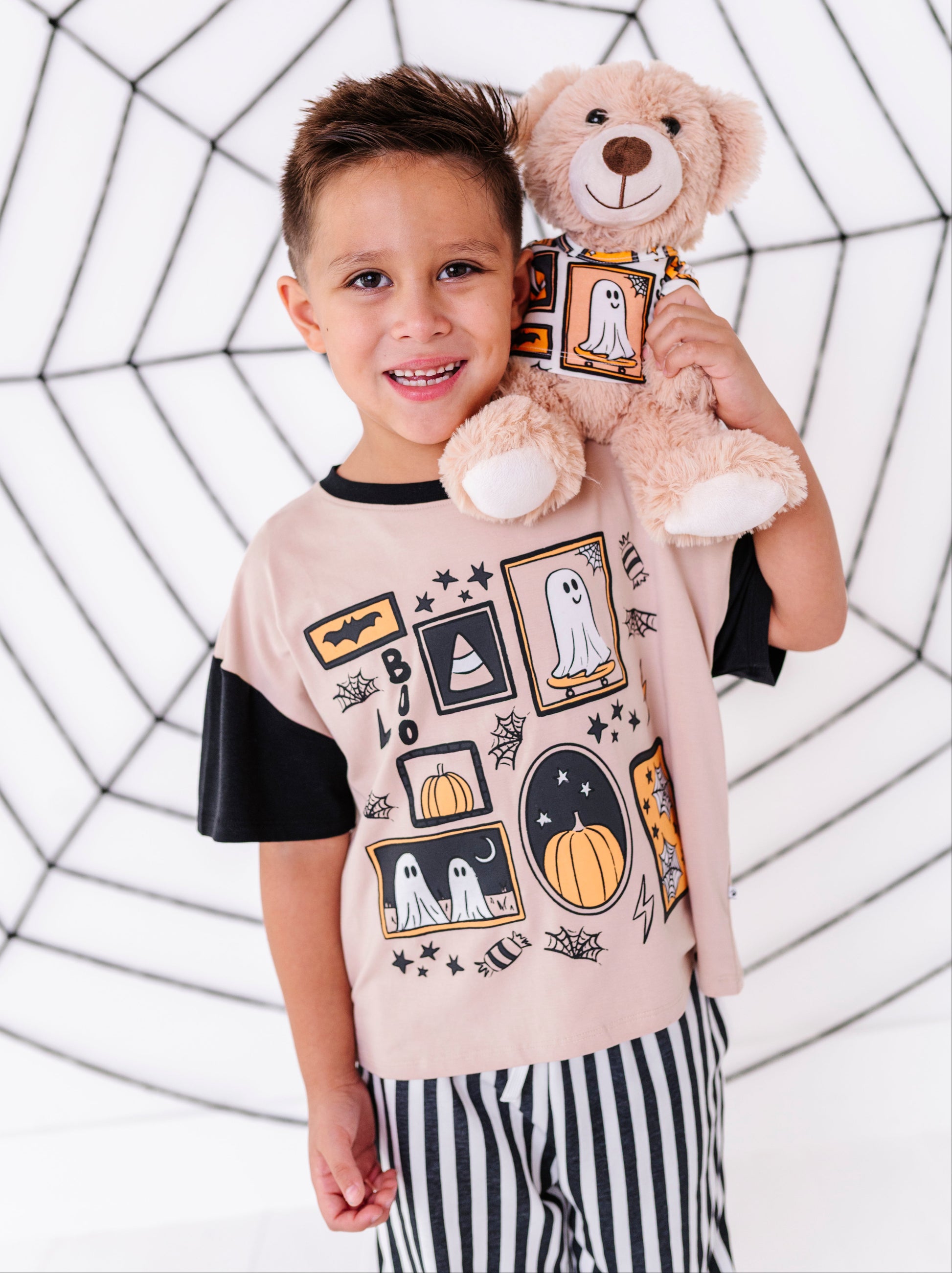 A young boy wearing a light tan and black short-sleeve t-shirt with a Halloween-themed graphic print, holding a plush teddy bear. The background is a white studio backdrop decorated with a black spiderweb pattern.