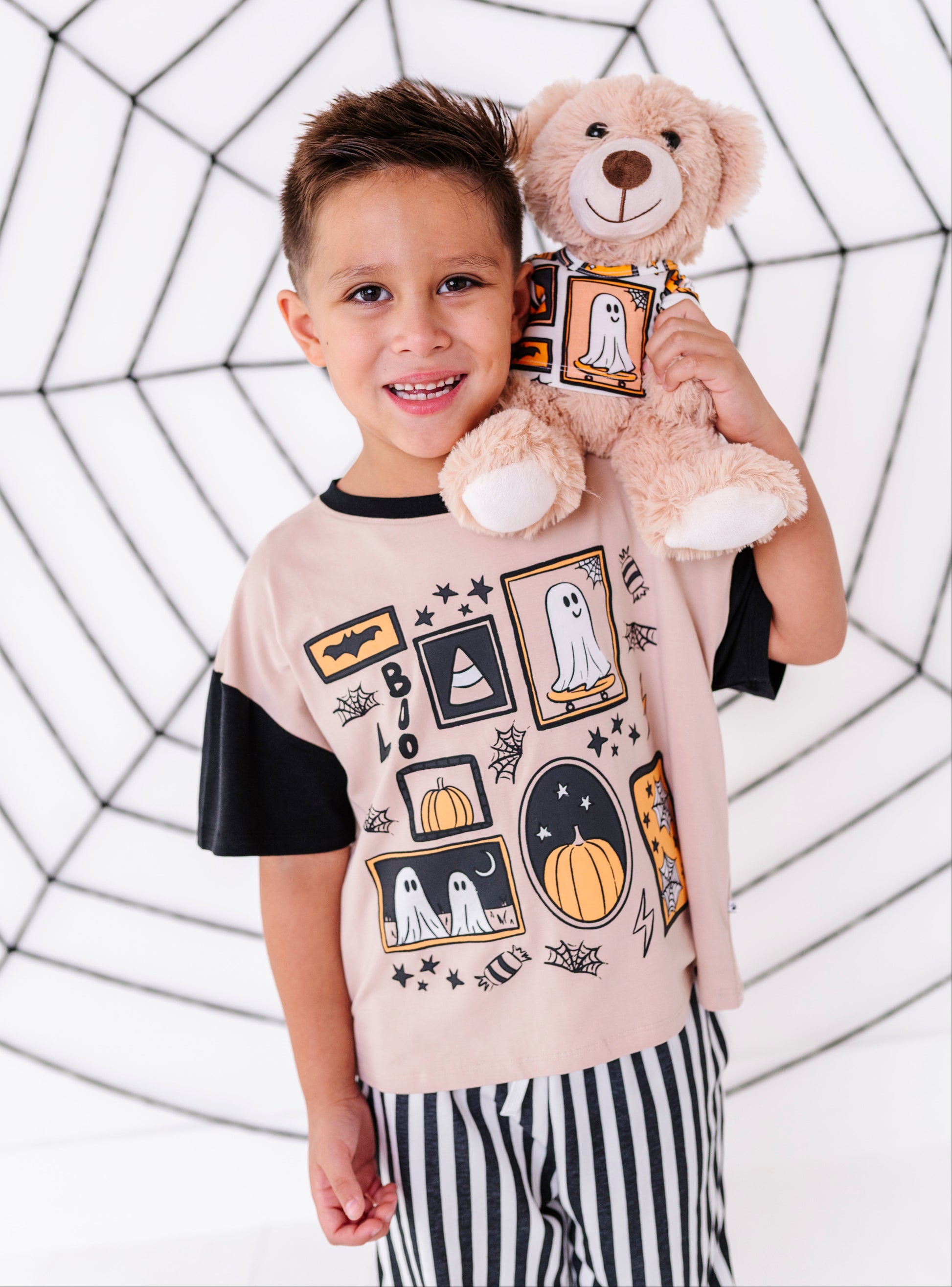 A young boy wearing a light pink and black short-sleeve t-shirt with a Halloween-themed graphic print, holding a plush teddy bear. The background is a white studio backdrop decorated with a black spiderweb pattern.