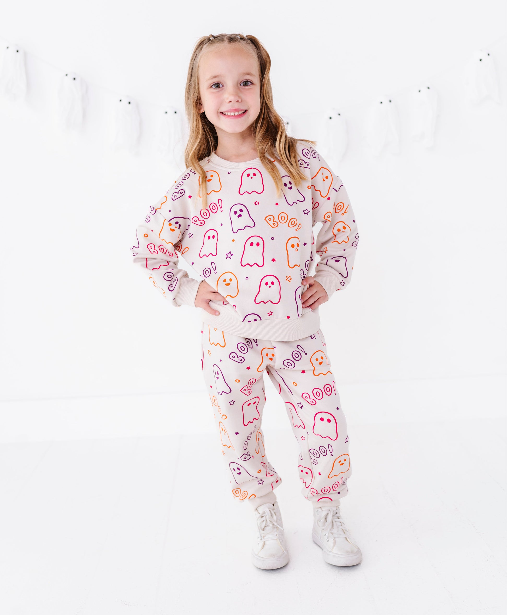 A young girl stands in a white studio wearing a matching two-piece pajama set with a white base and a colorful pattern of cartoon ghosts and hearts. She is wearing white sneakers.