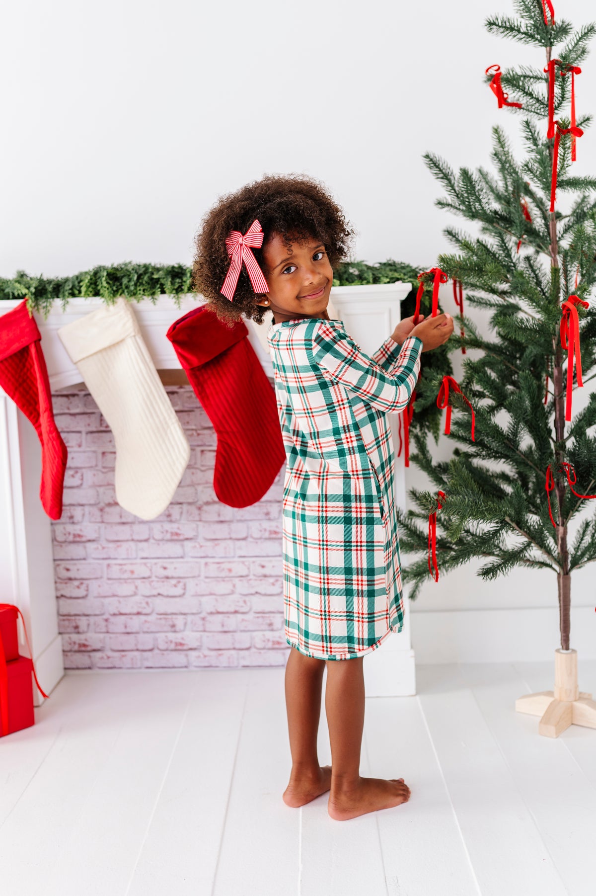 A little girl wearing a bamboo nightgown in a classic Christmas plaid print, decorating a Christmas tree