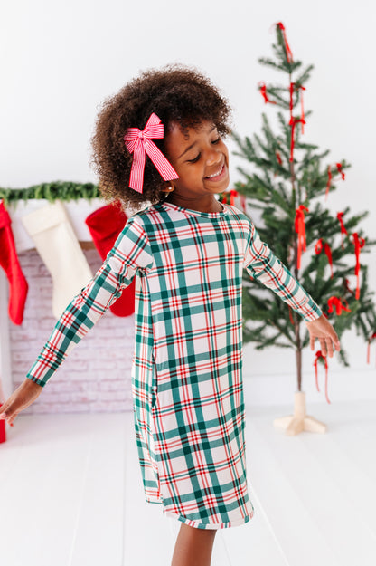 A little girl wearing a bamboo nightgown in a classic Christmas plaid print, standing in front of a fireplace with stockings and a decorated Christmas tree