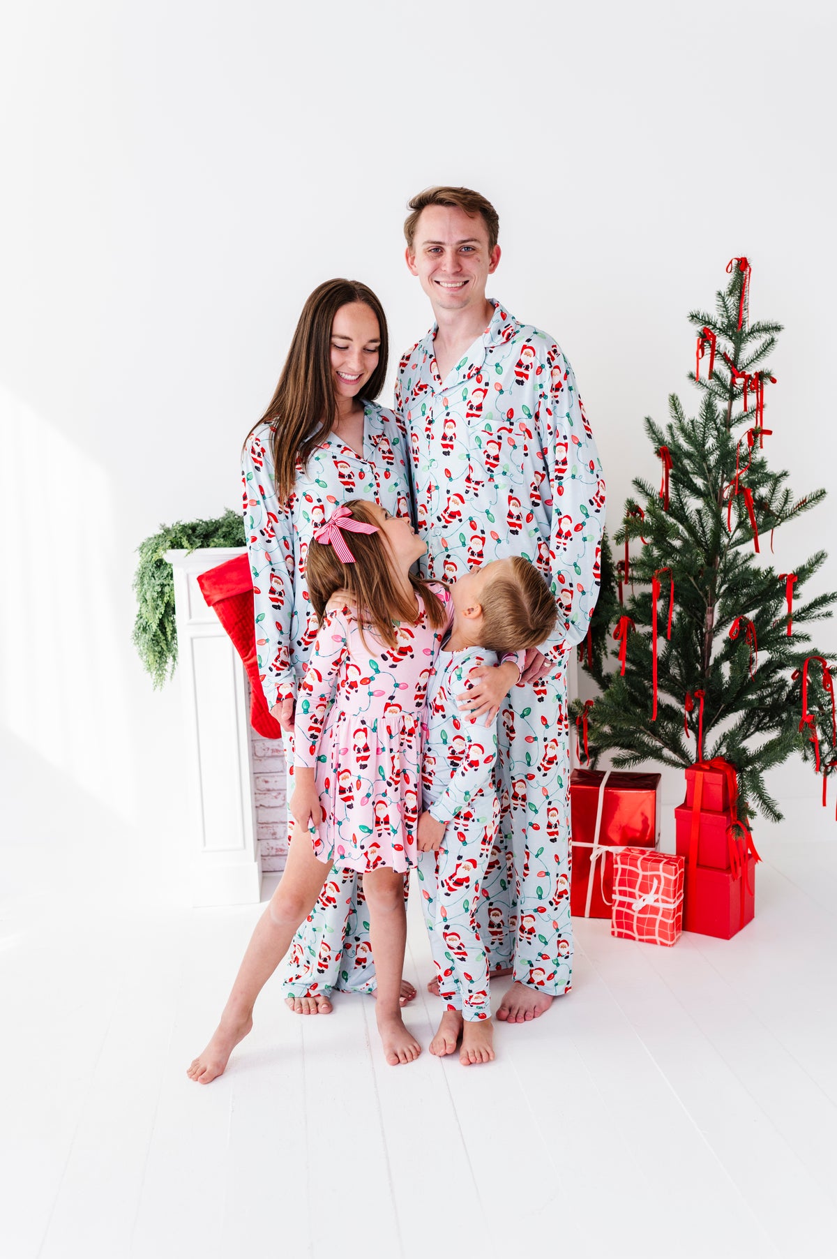 A family of four wearing matching Santa Claus bamboo pajamas poses together in a bright white room next to a decorated Christmas tree and wrapped gifts.