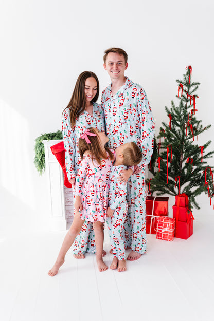 A family of four wearing matching Santa Claus bamboo pajamas poses together in a bright white room next to a decorated Christmas tree and wrapped gifts.