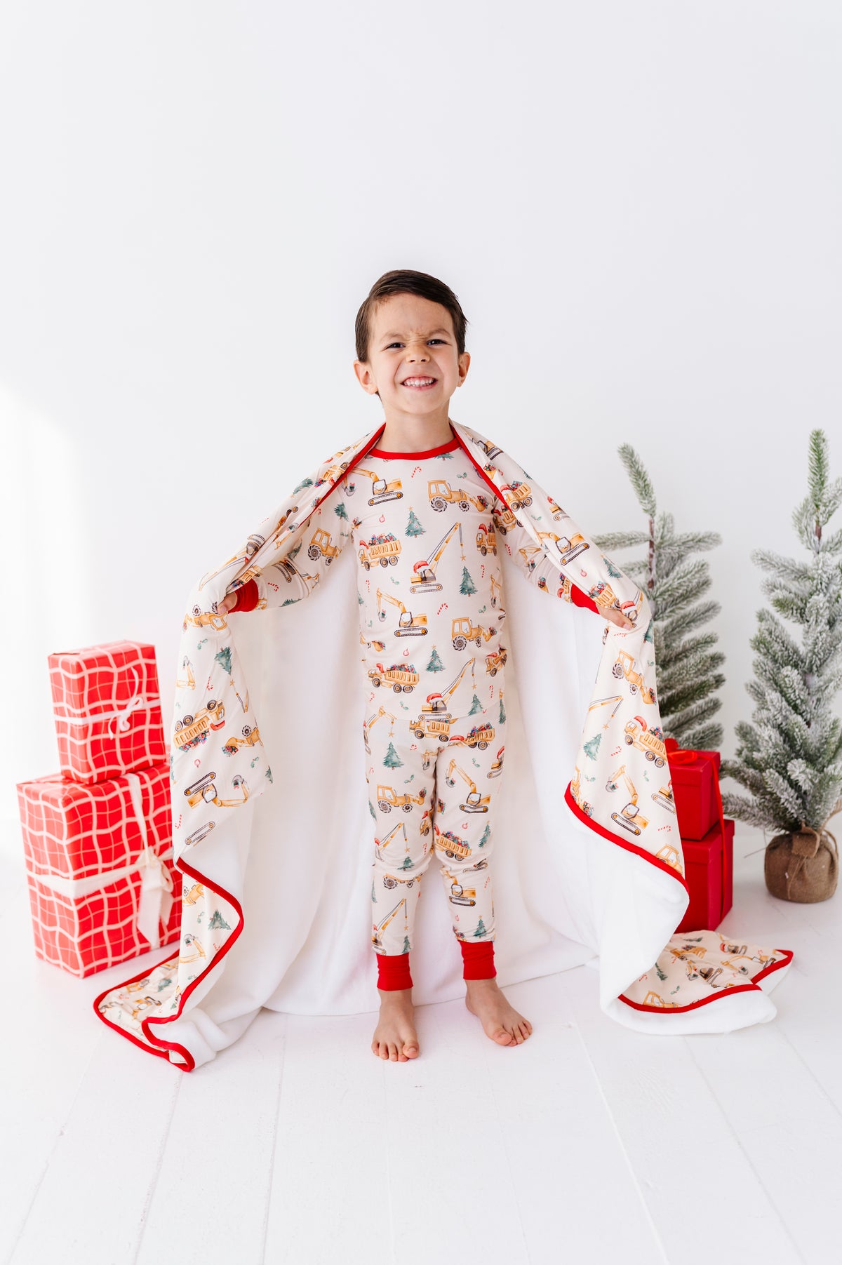 A young boy wearing festive Christmas-themed bamboo pajamas standing in a bright white room with trees and presents, wrapped in a matching construction-themed plush blanket 