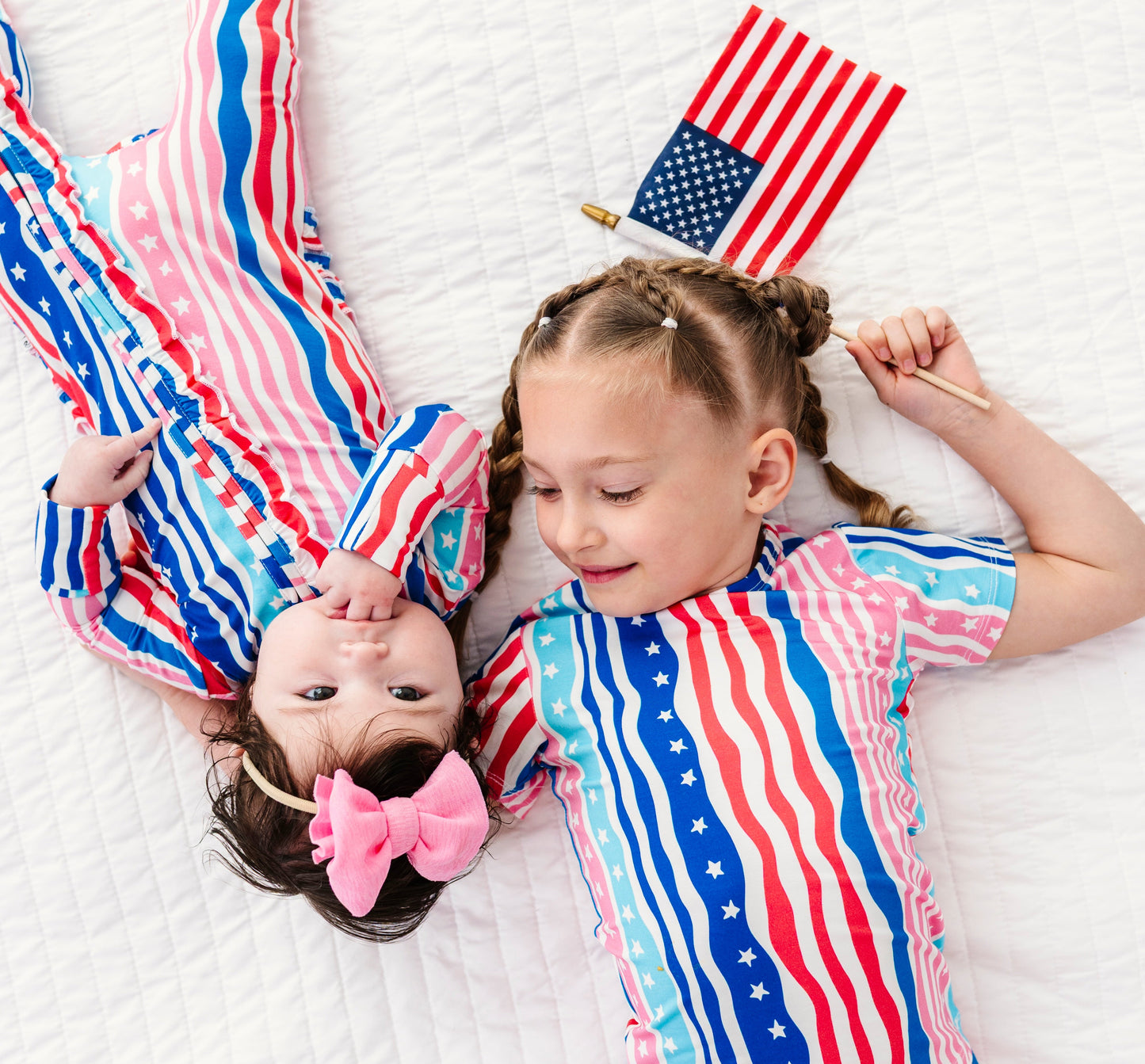 Two children in matching American flag-themed bamboo pajamas lying on a white blanket with small American flags.