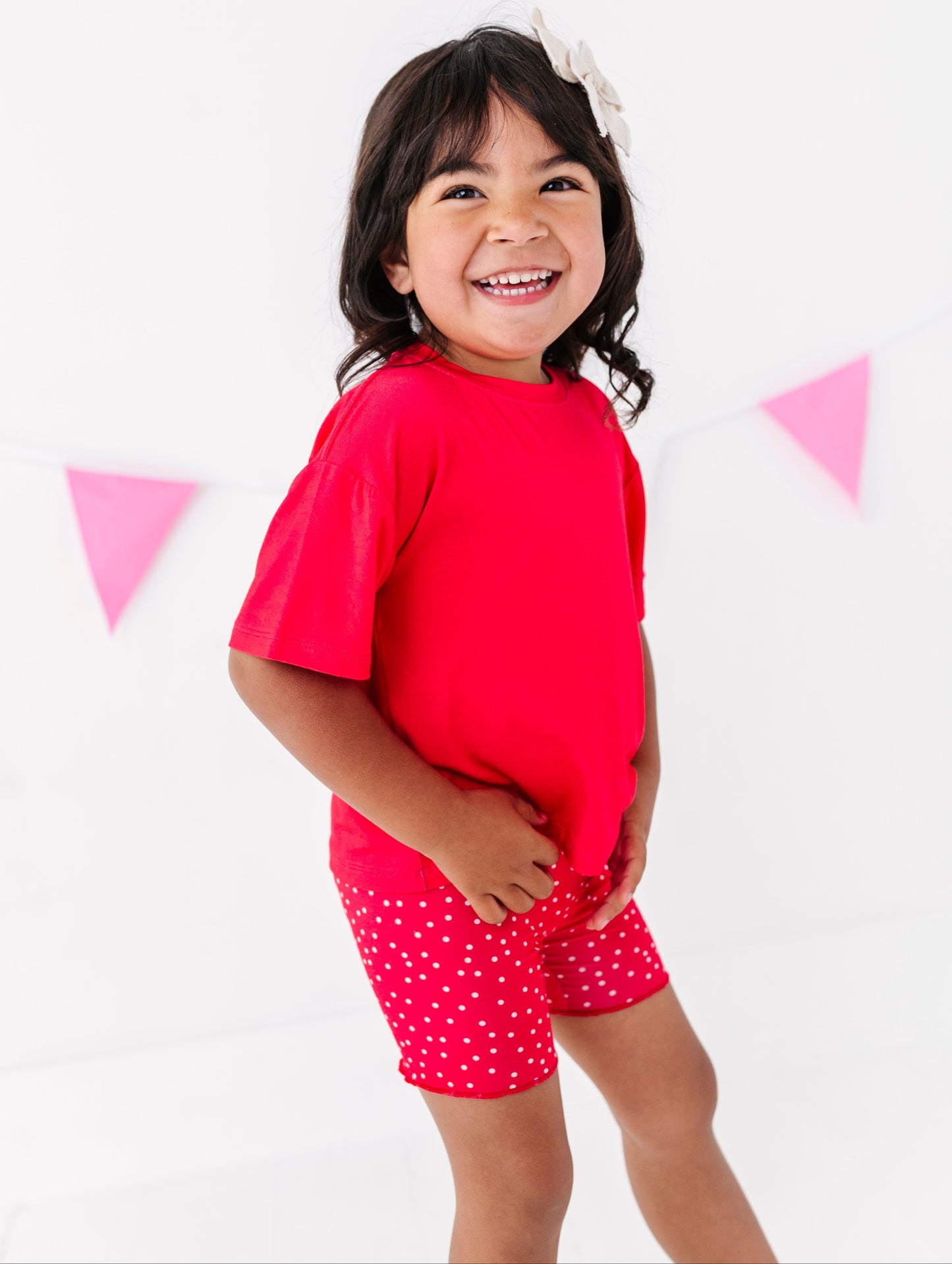 Child wearing a red shirt and red shorts with white polka dots, standing against a white background with pink triangle flags.