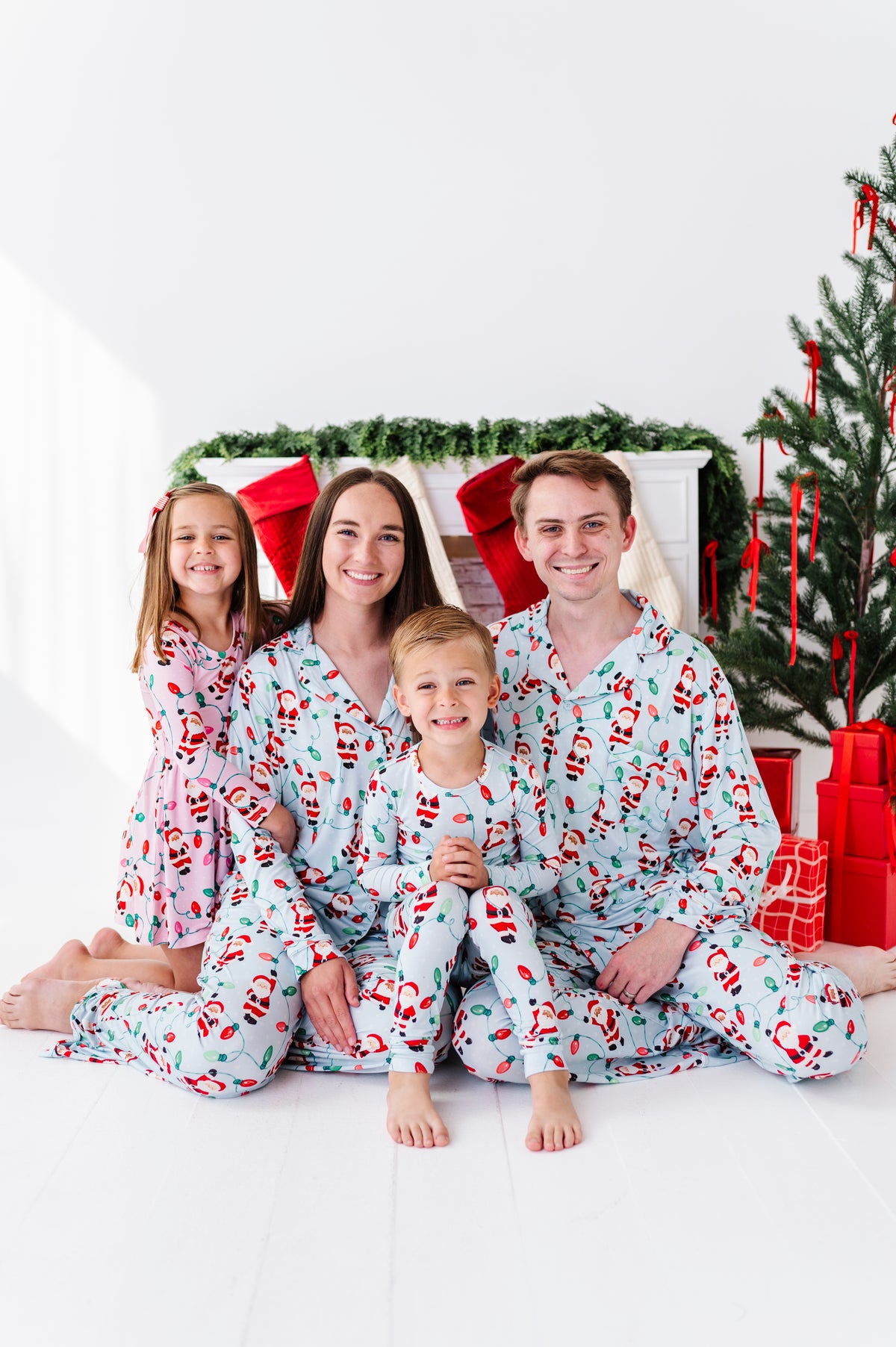 A family of four wearing matching light blue Christmas bamboo pajamas, sitting together in front of a decorated Christmas tree and fireplace.