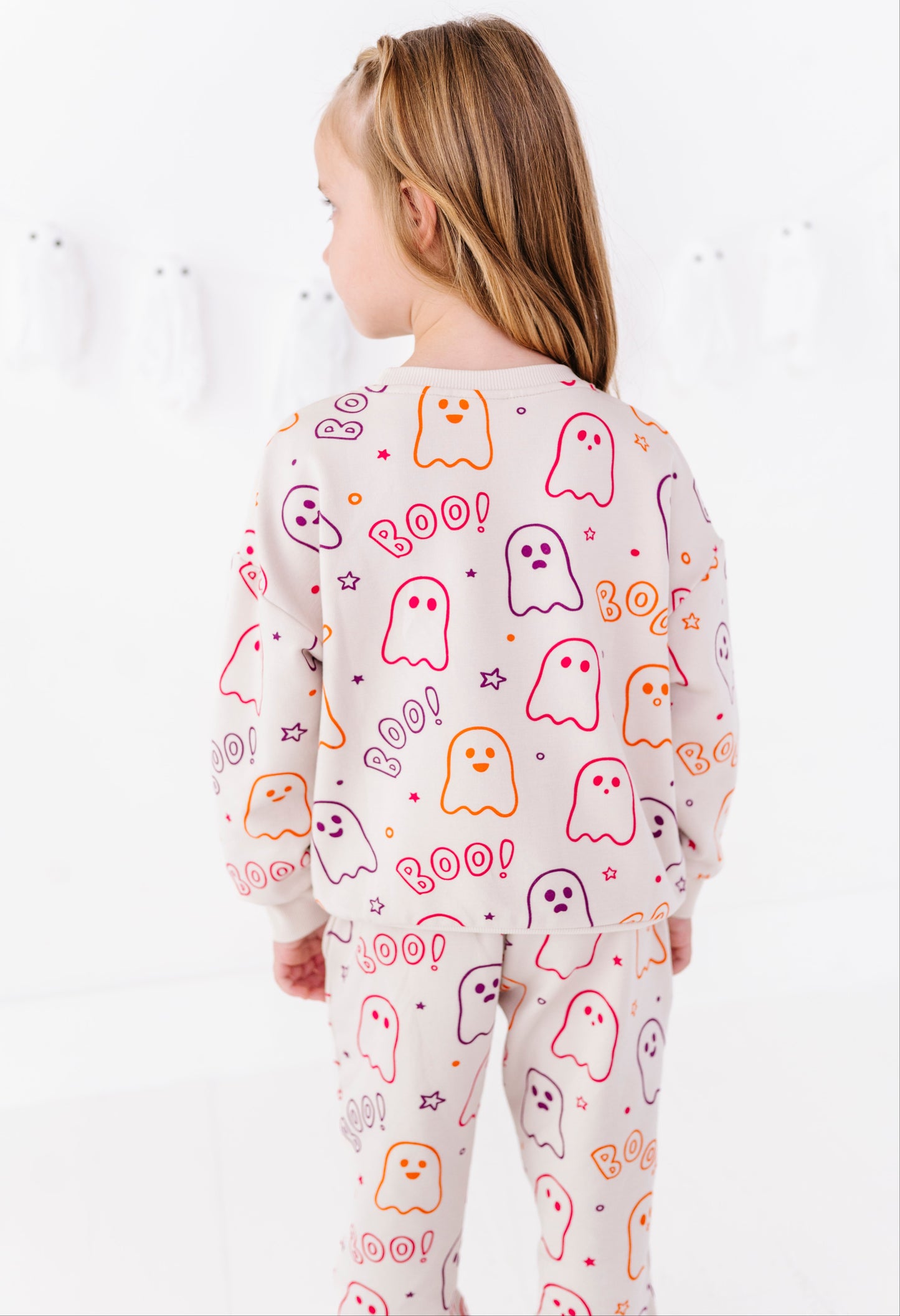 A young girl viewed from behind wearing a matching long-sleeve pajama set with a cute ghost pattern. The pajamas are cream-colored with colorful printed ghosts in pink, orange, and purple, along with 'BOO!' text.