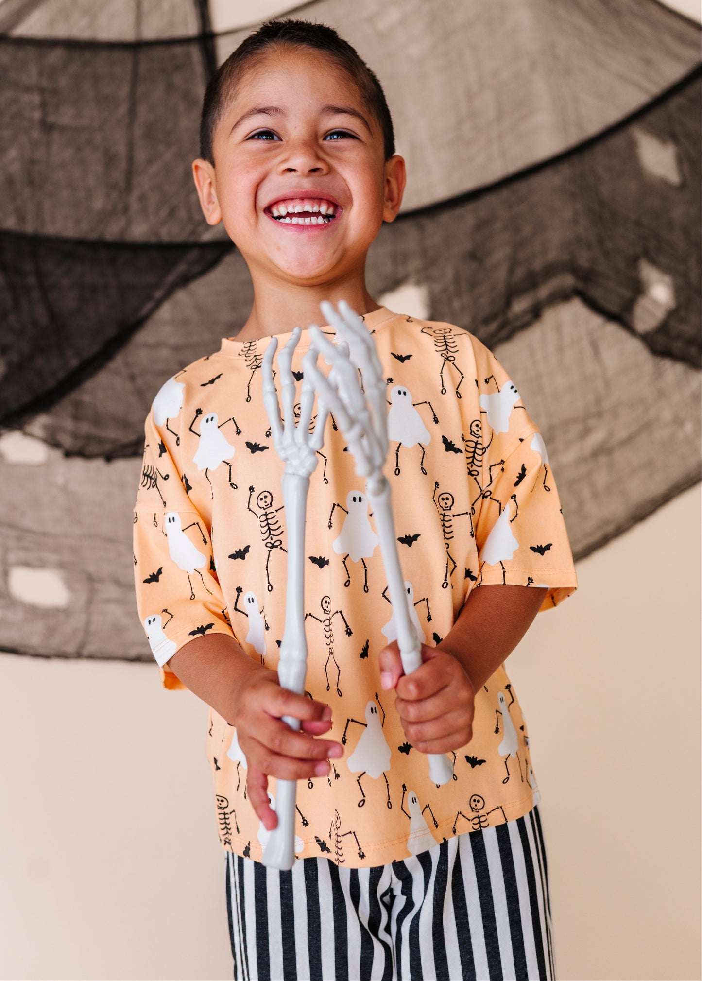 Child wearing a Halloween-themed shirt holding skeleton hands against a decorative backdrop.