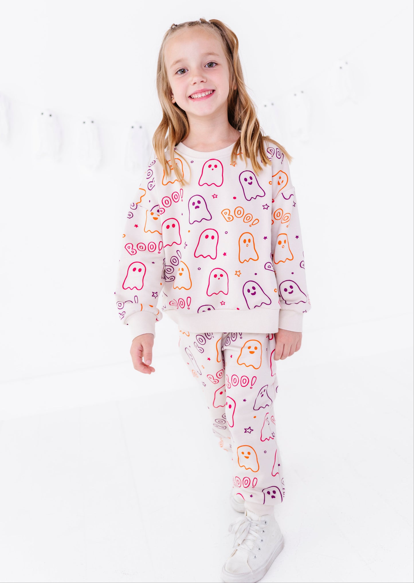 A young girl with blonde pigtails stands against a white studio background wearing a matching two-piece pajama set. The set features a light cream sweatshirt and pants covered in a playful pattern of colorful ghosts and the word 'BOO!'.
