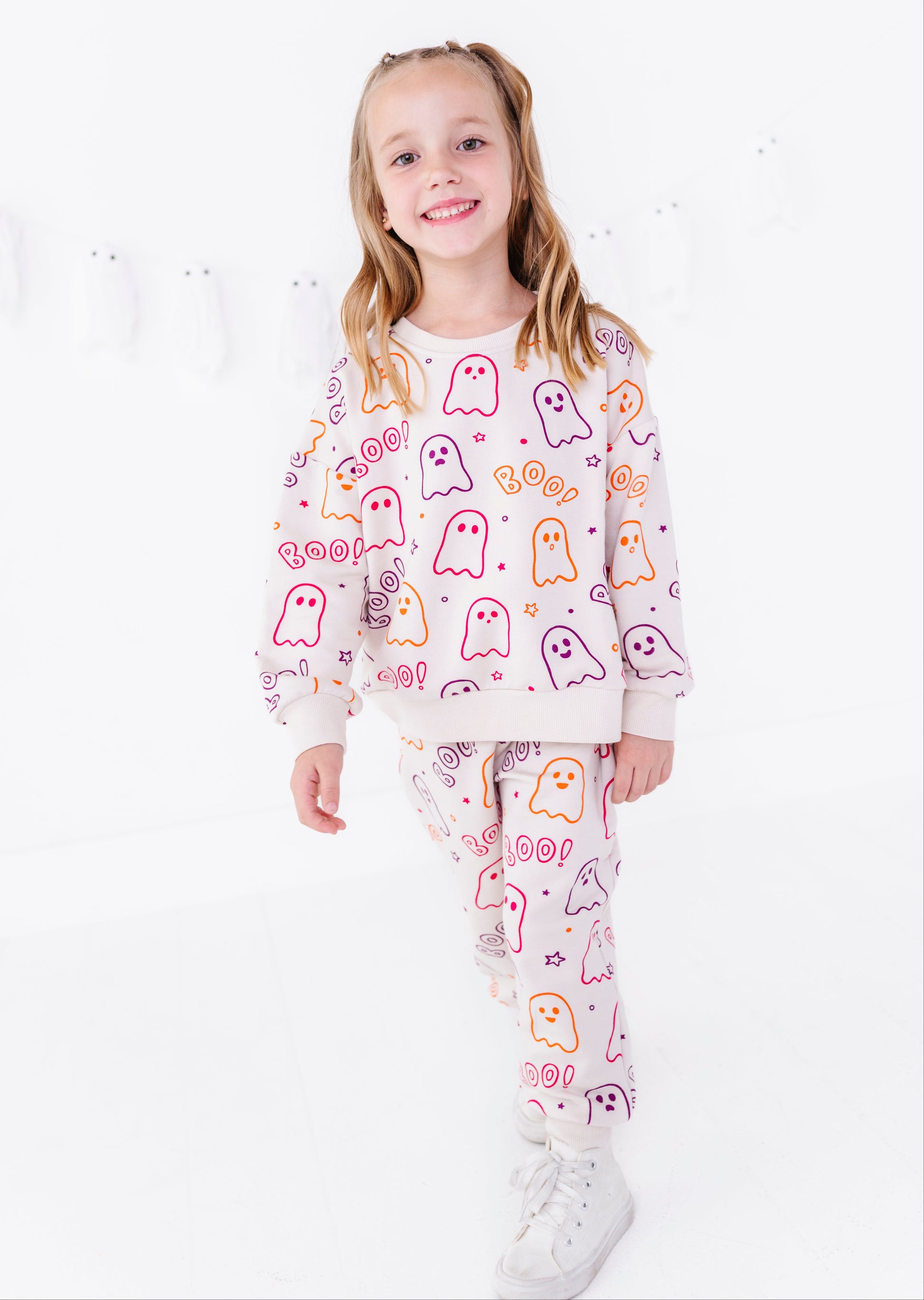 A young girl with blonde pigtails stands against a white studio background wearing a matching two-piece pajama set. The set features a light cream sweatshirt and pants covered in a playful pattern of colorful ghosts and the word 'BOO!'.