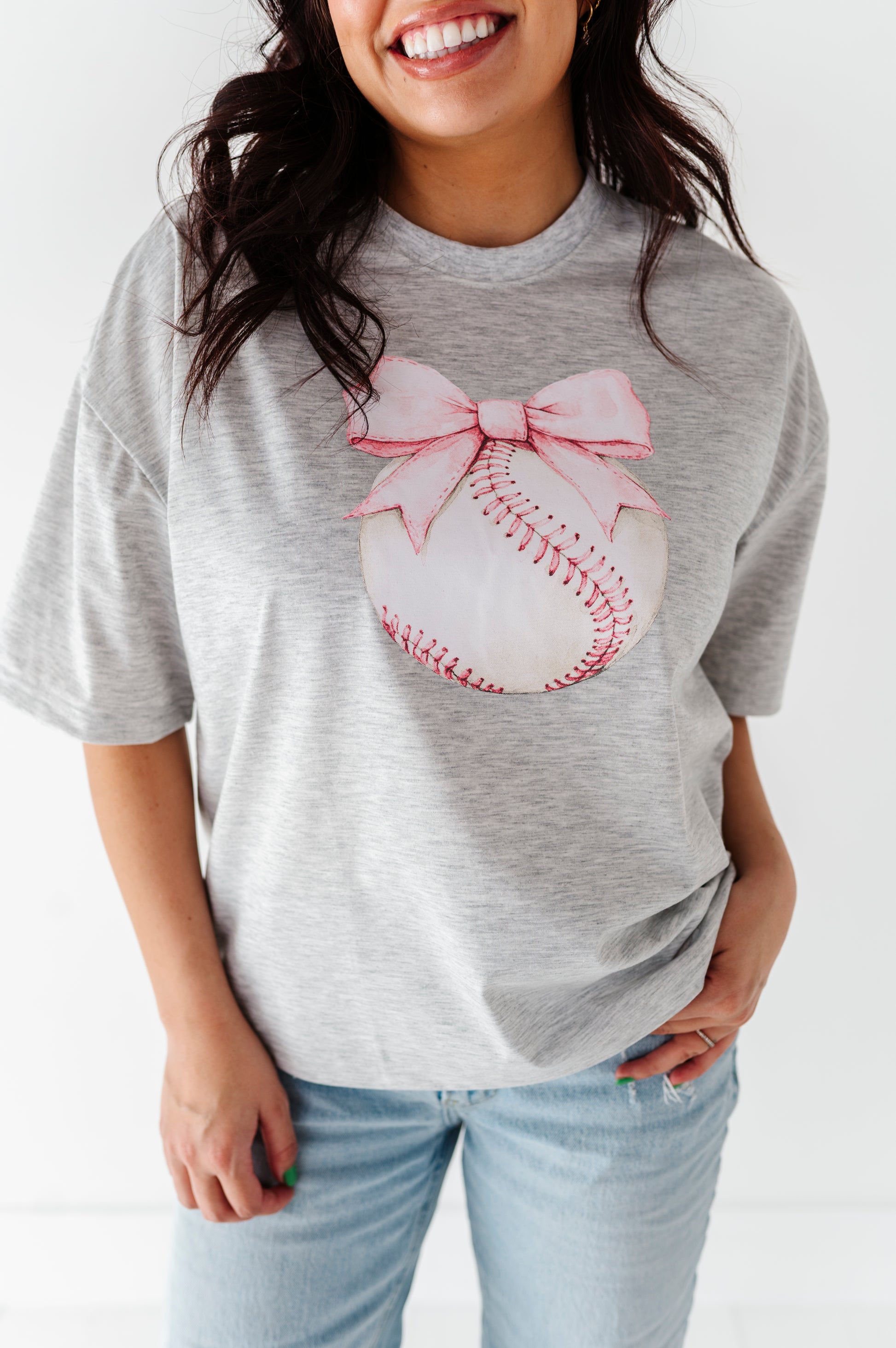 Mom wearing a gray t-shirt with a baseball and pink bow design, smiling against a white background