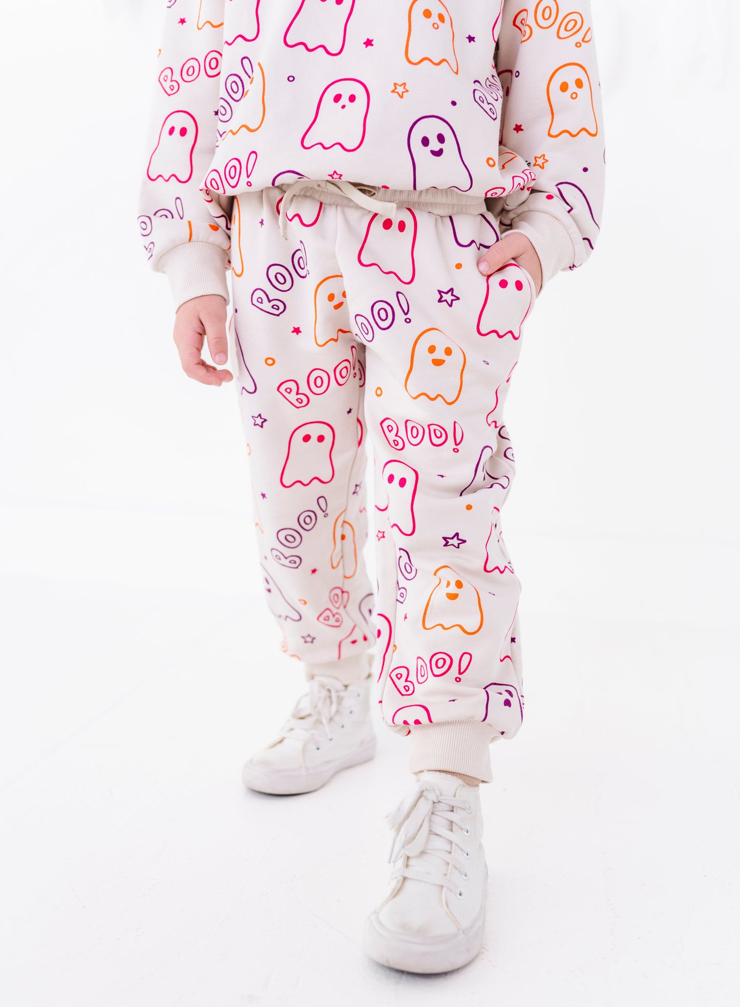 A child wearing a matching light pink sweatshirt and jogger pants set with a colorful ghost and 'Boo! Roar!' Halloween print, paired with white sneakers, against a plain white background.