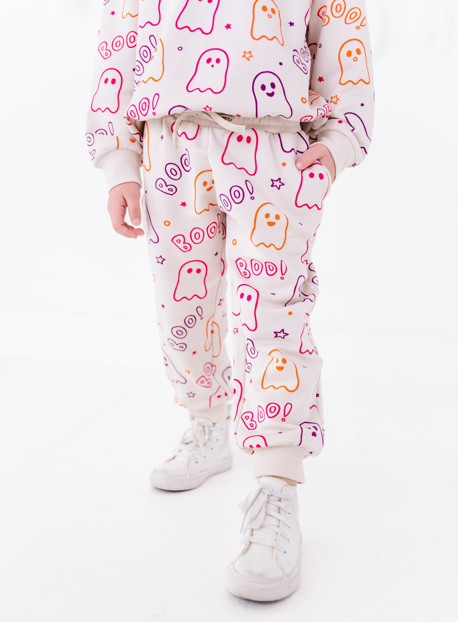 A child wearing a matching light pink sweatshirt and jogger pants set with a colorful ghost and 'Boo! Roar!' Halloween print, paired with white sneakers, against a plain white background.