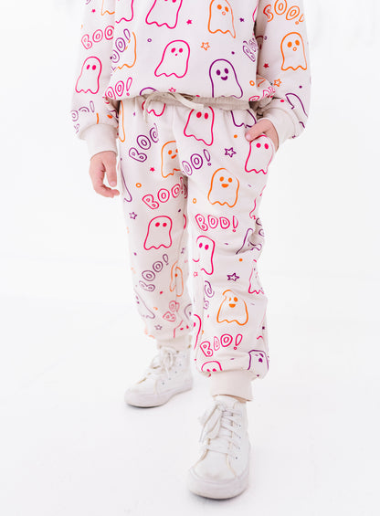 A child wearing a matching light pink sweatshirt and jogger pants set with a colorful ghost and 'Boo! Roar!' Halloween print, paired with white sneakers, against a plain white background.
