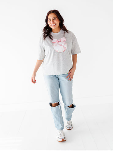 Woman wearing a gray t-shirt with a baseball and bows graphic design and light blue jeans on a white background