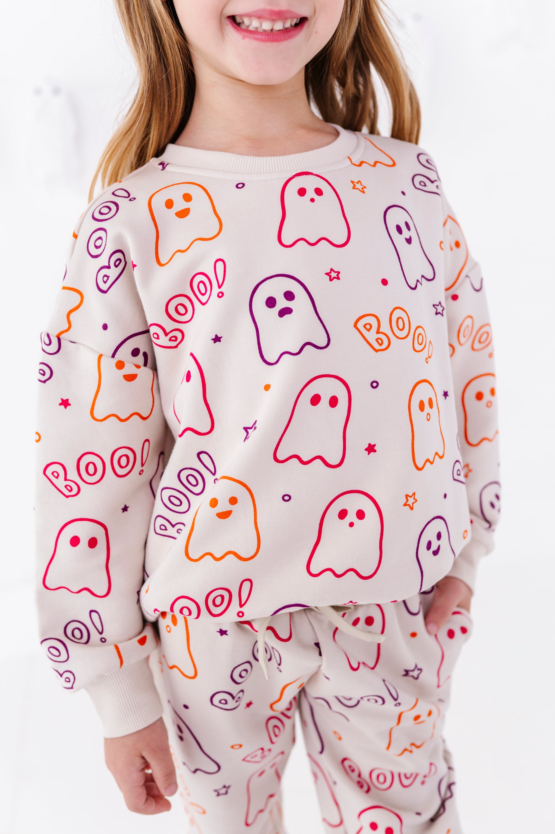 A child wearing a matching long-sleeve sweatshirt and sweatpants set with a playful ghost pattern in orange, purple, and pink on a cream-colored fabric, set against a bright white background.